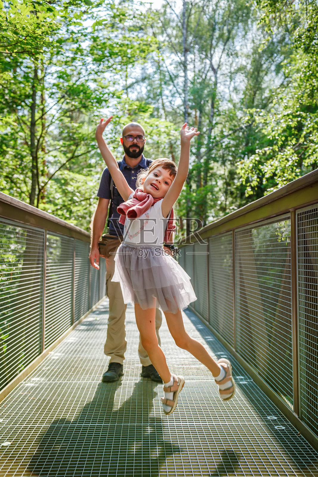 小女孩在森林桥上快乐地跳跃,表达着兴奋和快乐。她的父亲在背景中微笑。照片摄影图片
