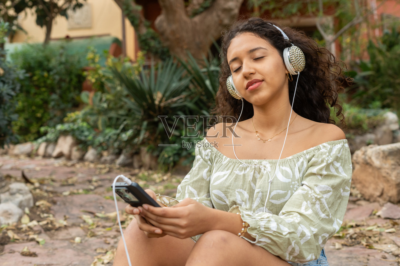 年轻女性坐在宁静的花园中,戴着无线耳机享受音乐,闭眼放松。照片摄影图片