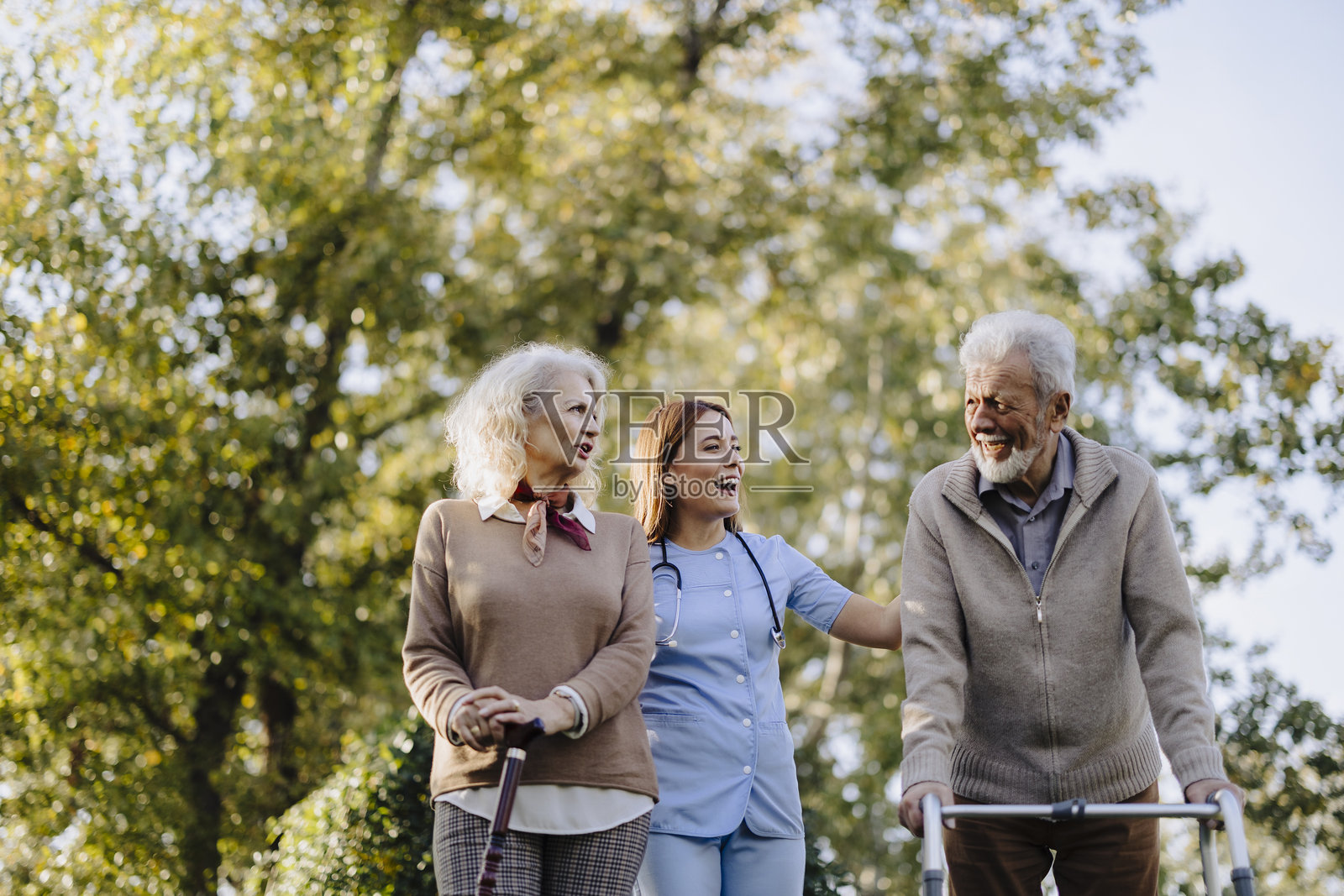 护士与老年夫妇在花园中散步,提供帮助和支持,享受关怀。养老院,微笑的退休人员与护理人员在医疗、康复和锻炼中快乐地互动。照片摄影图片