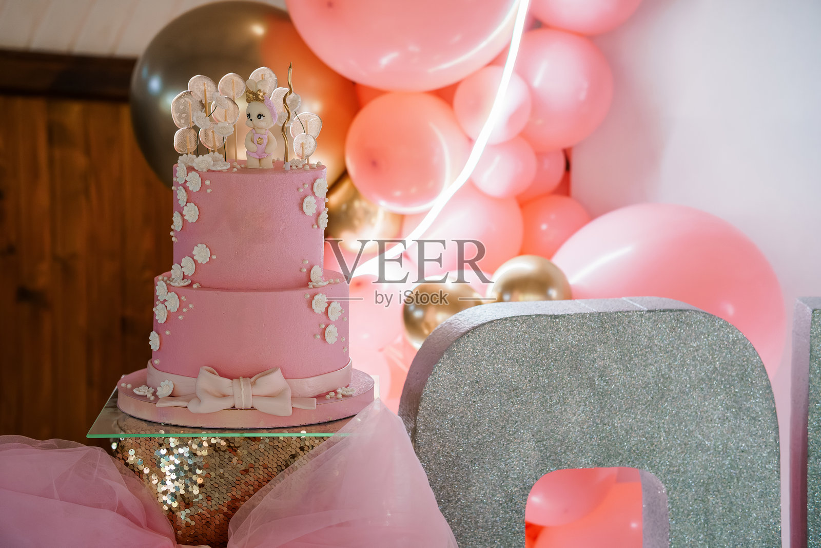 可爱的双层粉色小女孩一岁生日蛋糕,配有大象装饰,背景是节日气球布置。照片摄影图片