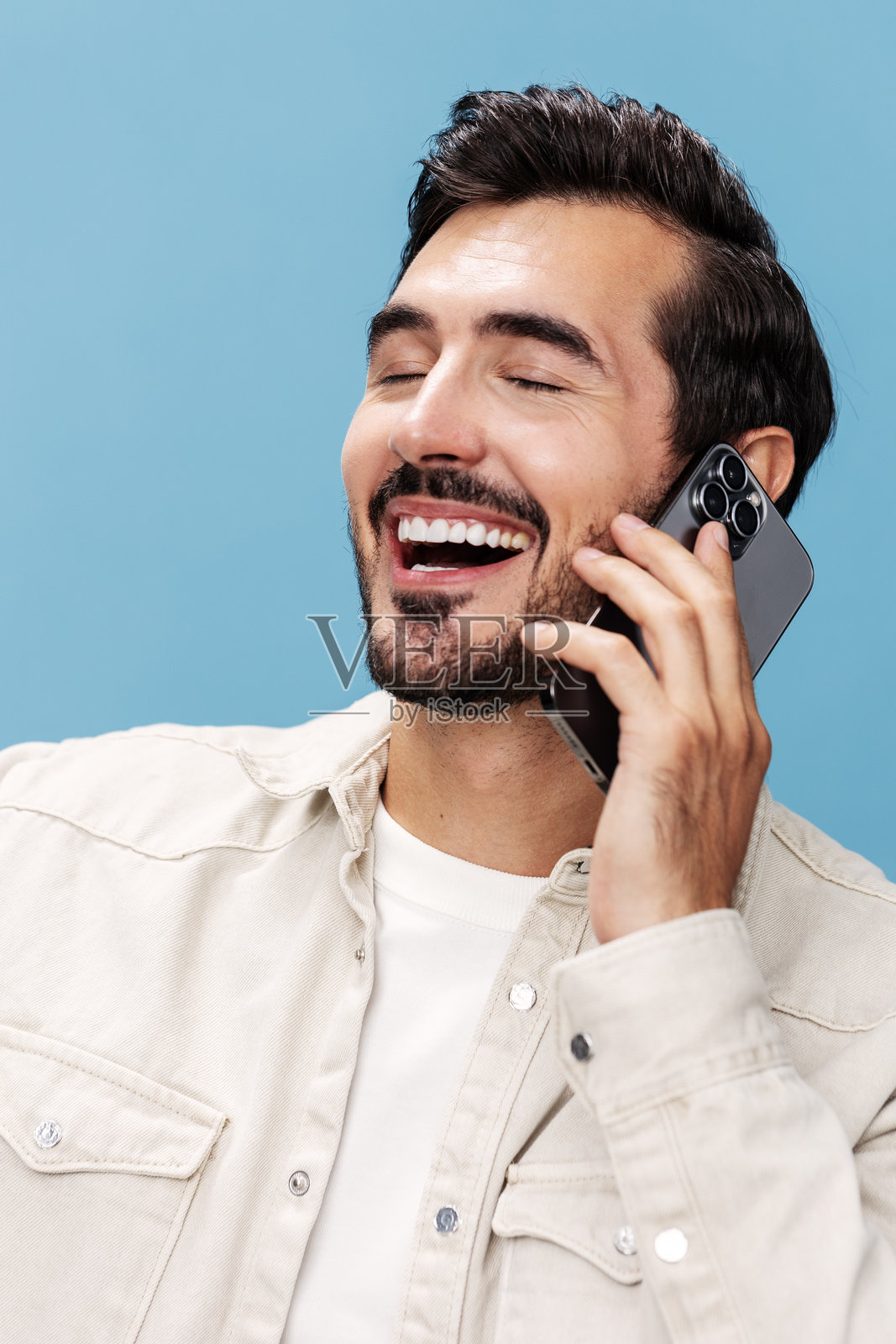 棕发男子在蓝色背景下穿着白色T恤和牛仔裤,微笑着打电话,露出牙齿,充满快乐和笑声,留有空白空间。照片摄影图片