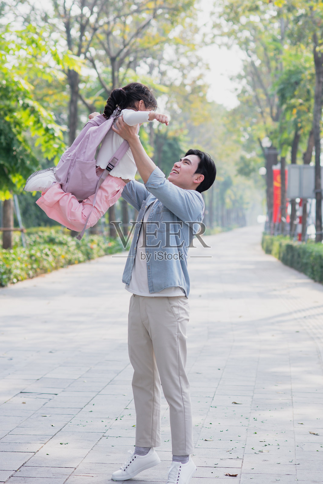 父亲在阳光明媚的公园中快乐地举起女儿,展现出浓浓的爱意和顽皮的气息,四周绿树成荫。照片摄影图片