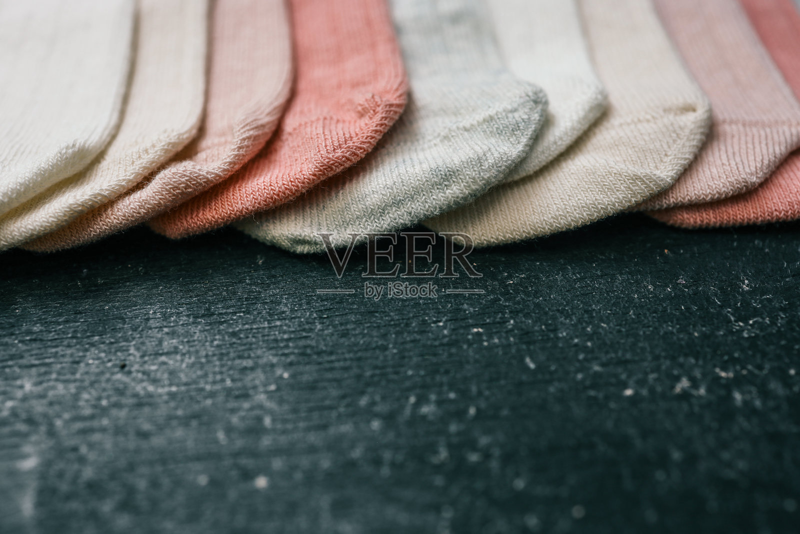 一排柔软的婴儿袜子,颜色为白色、桃色、珊瑚色、米色和浅灰色,整齐地排列在深色纹理表面上。舒适、简约的平铺设计,适合婴儿服装产品展示。照片摄影图片