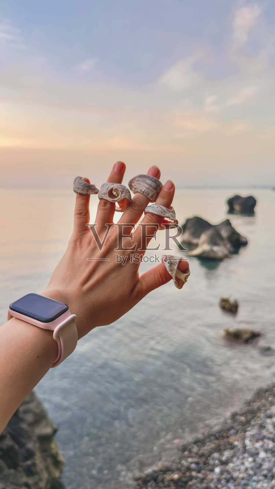 一只装饰着贝壳如同戒指的女人手在日落时分高举于平静的海面前。水中耸立着岩石,天空被柔和的晚霞渲染成粉彩色调。垂直旅行照片照片摄影图片