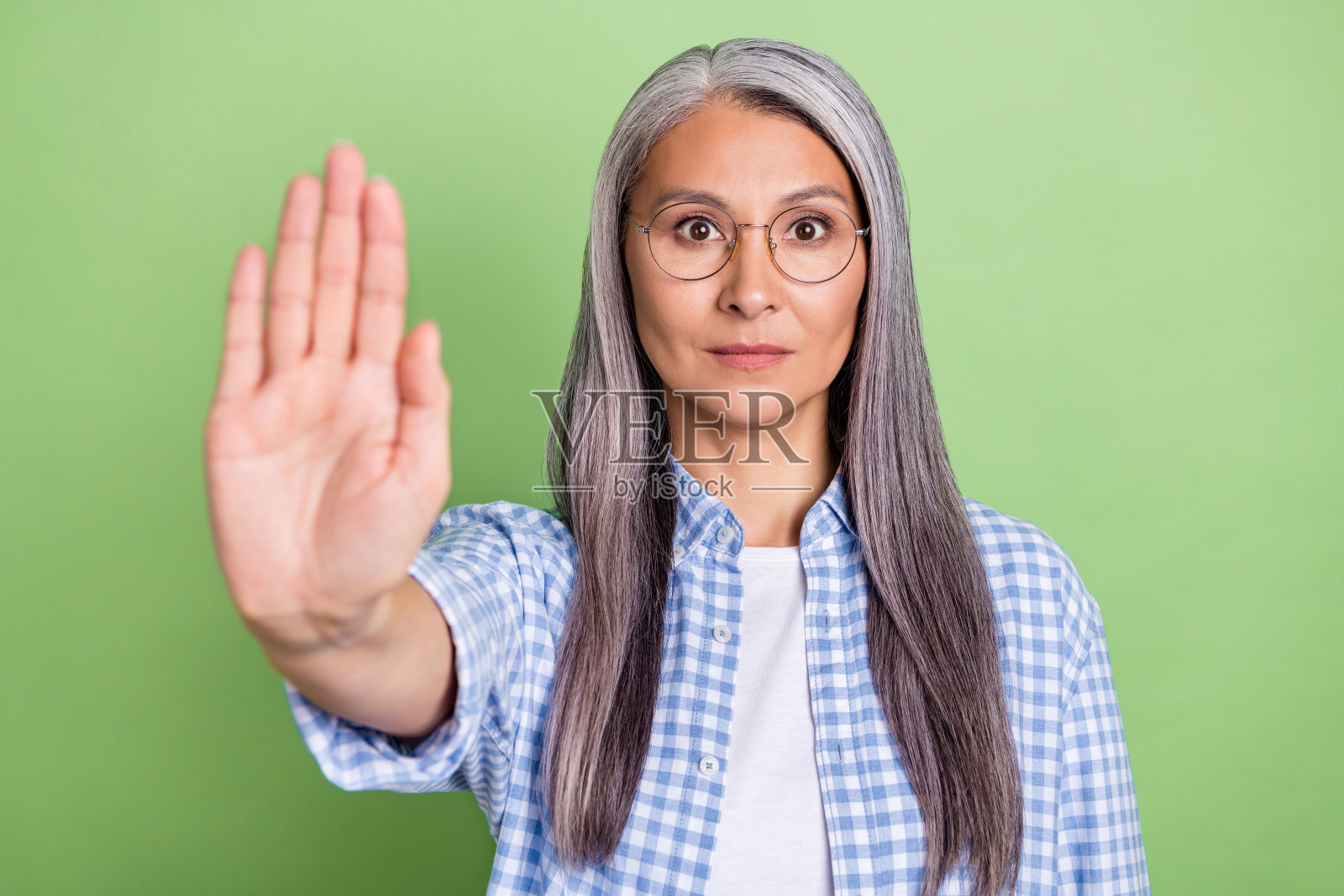 一位严肃的灰发女士,戴着眼镜,穿着格子衬衫,站在绿色背景前。照片摄影图片