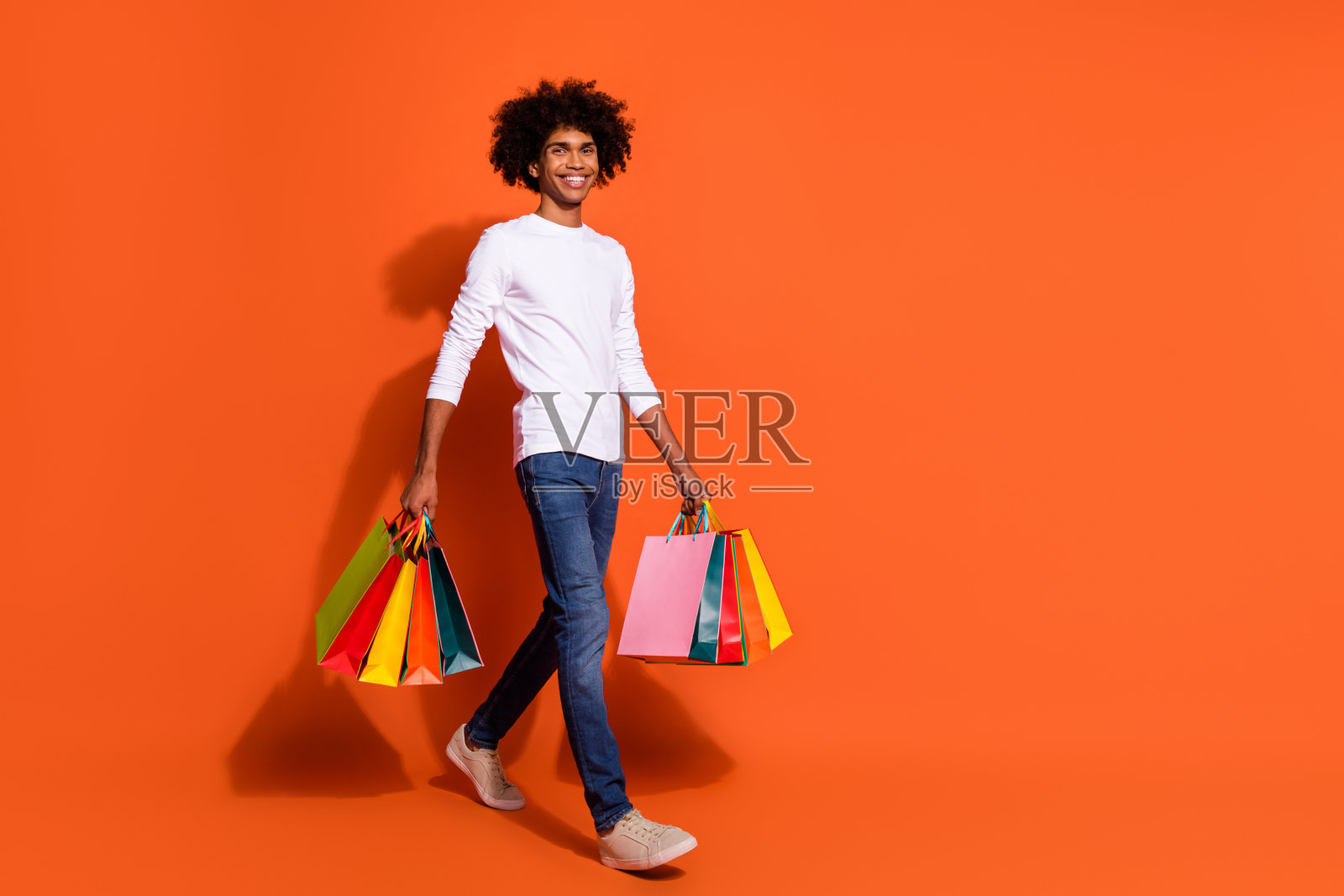 穿着白衬衫的帅气小伙子在橙色背景下走在空旷的地方,手里提着购物袋。照片摄影图片