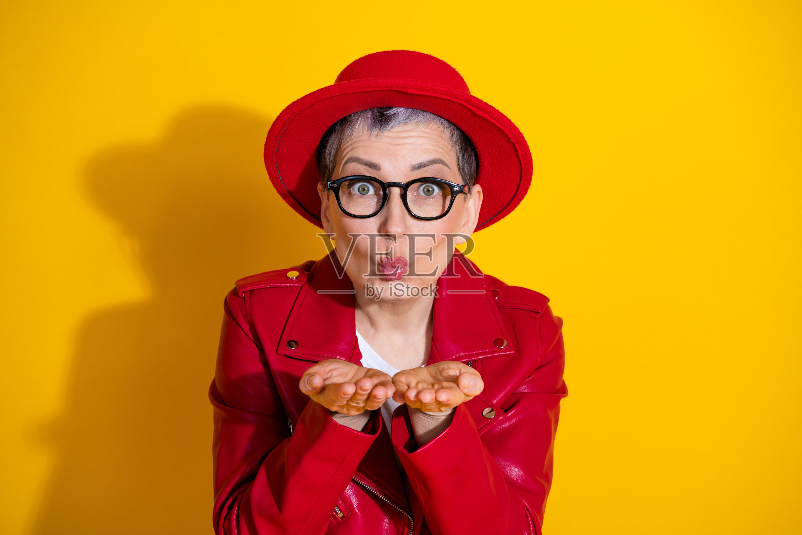 时尚的老年女性穿着红色皮夹克和帽子,手势表达,背景为明亮的黄色。照片摄影图片