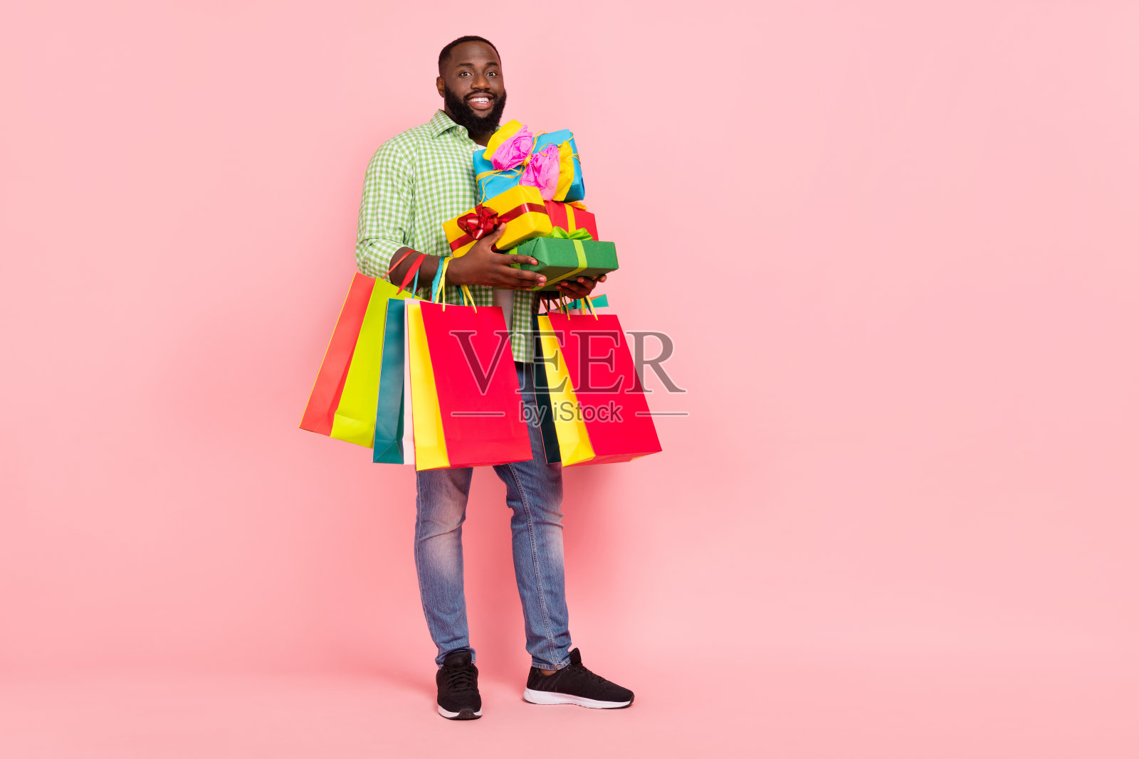 可爱年轻男孩手持礼物袋,穿着绿色衬衫、牛仔裤和运动鞋,背景为粉色。照片摄影图片