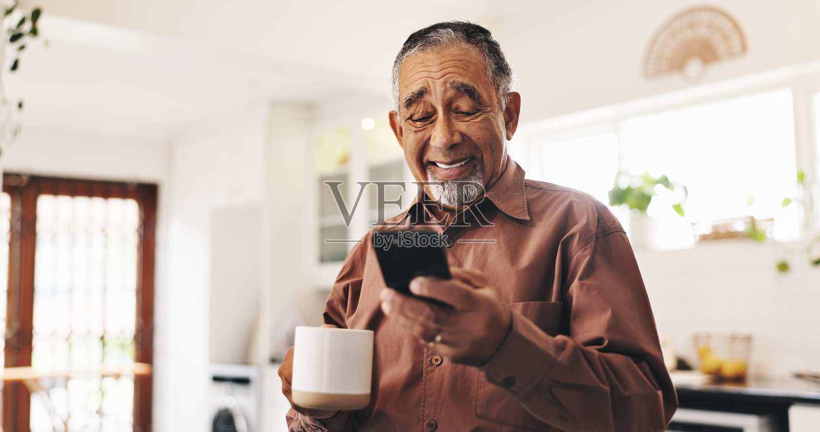 退休之家厨房里的老人,手握咖啡和手机,面带微笑,适合交流或发短信。快乐的老人在公寓中享受咖啡饮品,放松并使用社交媒体。照片摄影图片