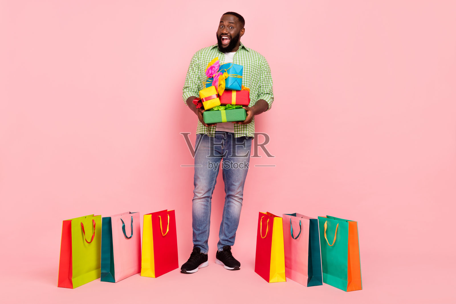 全身照,印象深刻的千禧一代男士手持礼物袋,穿着绿色衬衫和牛仔裤,脚穿运动鞋,背景为粉色。照片摄影图片