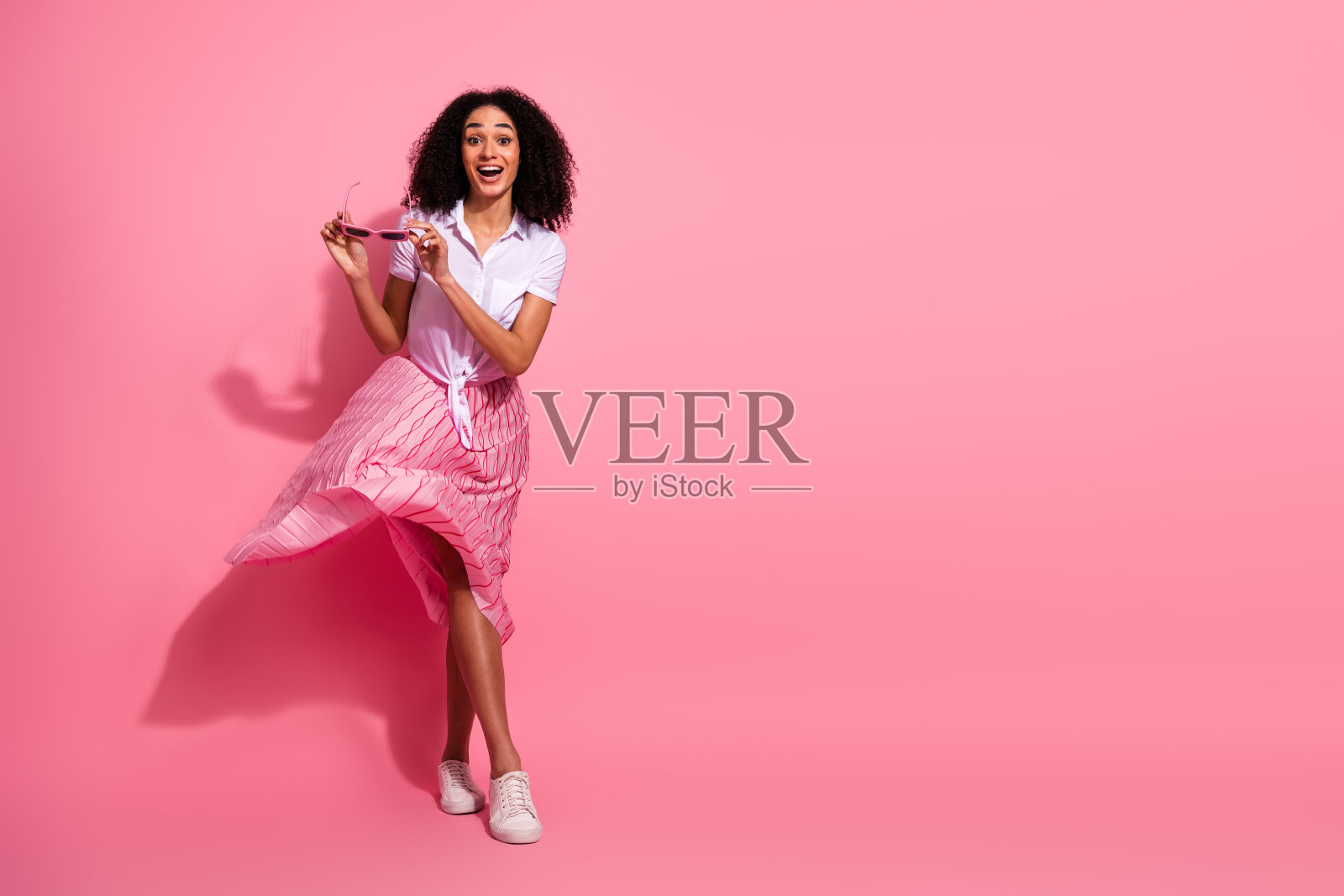 年轻时尚女性穿着休闲服,戴着太阳镜,自信地在鲜艳的粉色背景前摆姿势照片摄影图片