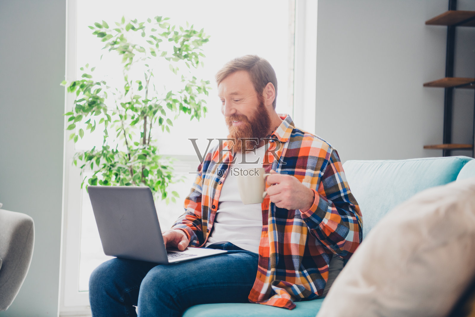 中年男子在周末的室内放松,手里拿着笔记本电脑和咖啡,享受着温馨客厅的闲暇时光。照片摄影图片