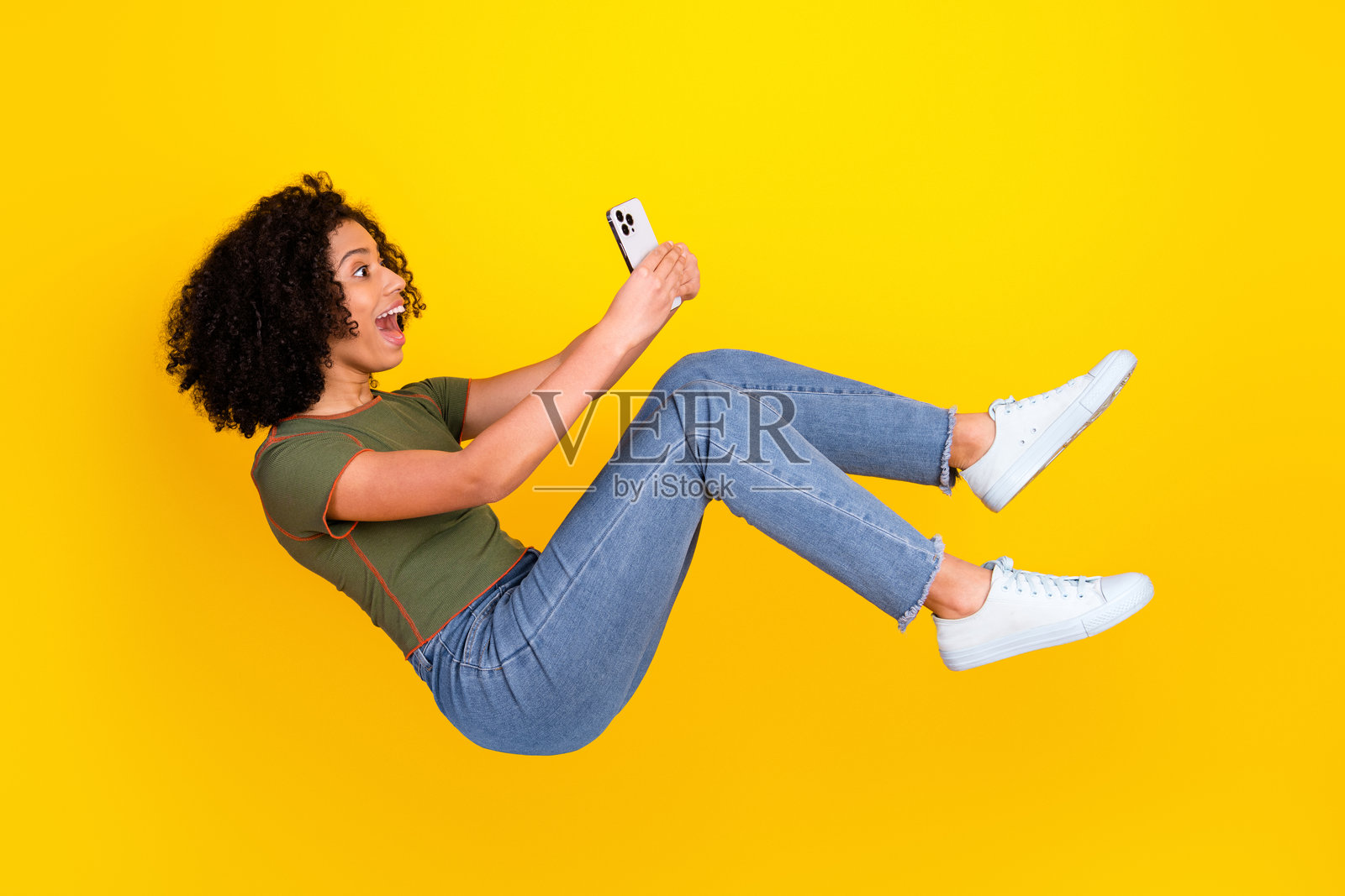 一位快乐的年轻女性,卷发,正看着她的智能手机,漂浮在鲜艳的黄色背景前。照片摄影图片