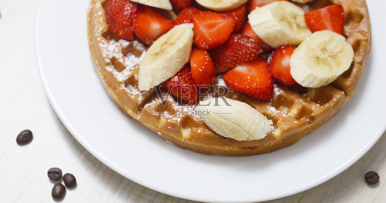 金黄酥脆的华夫饼,上面点缀着切片草莓和香蕉,撒上糖粉,静静地放在白色盘子上,旁边散落着咖啡豆,早晨的奢华享受。照片摄影图片