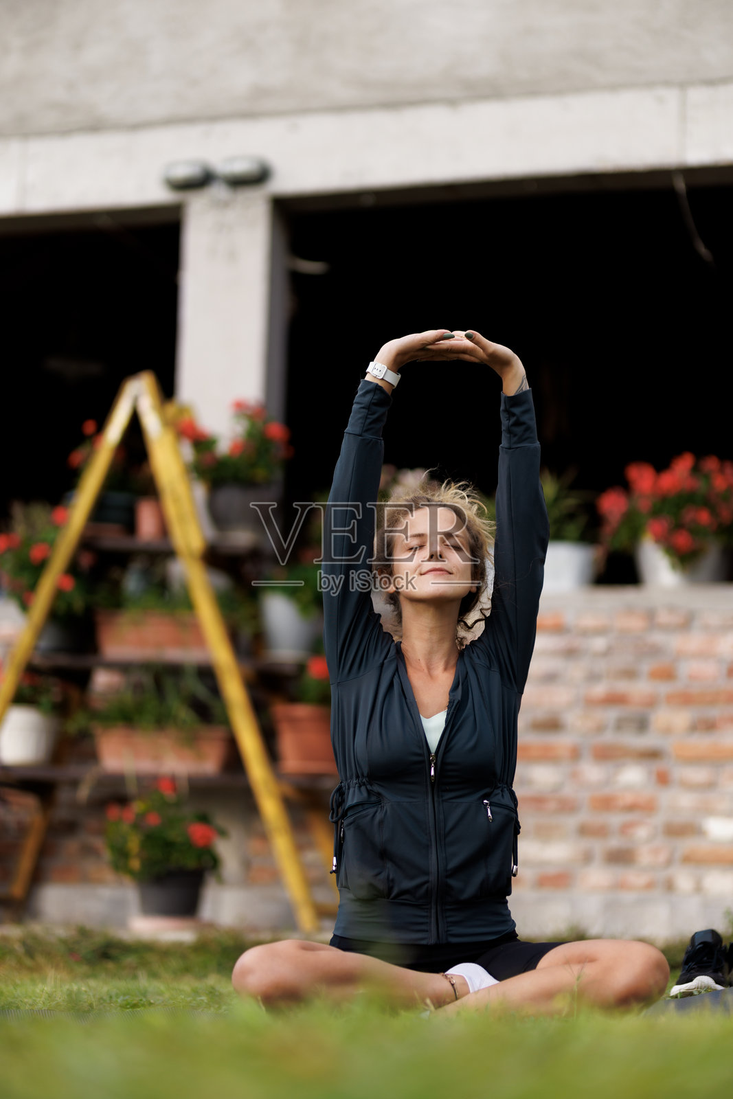 女性在花园环境中白天练习瑜伽,面带宁静的表情,伸展双臂。照片摄影图片