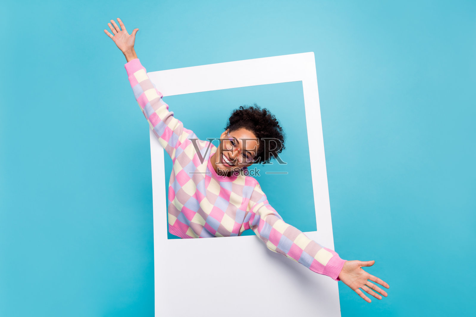 一个迷人快乐的女孩从照片框中走出来,向大家打招呼,享受乐趣,背景是明亮的蓝色。照片摄影图片