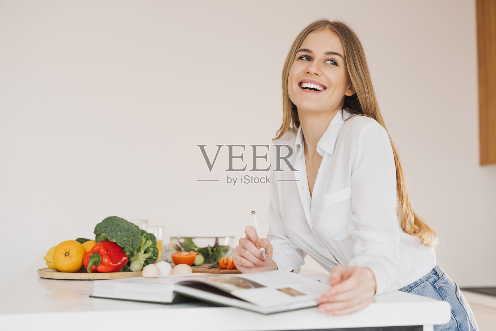 一位快乐可爱的金发女性在厨房里写东西,并查看食谱书。照片摄影图片