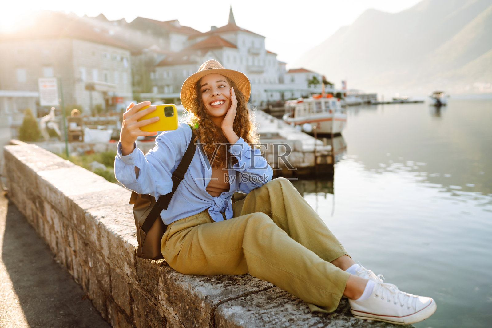 一位年轻的卷发女子戴着帽子,手持手机在水边拍自拍。照片摄影图片