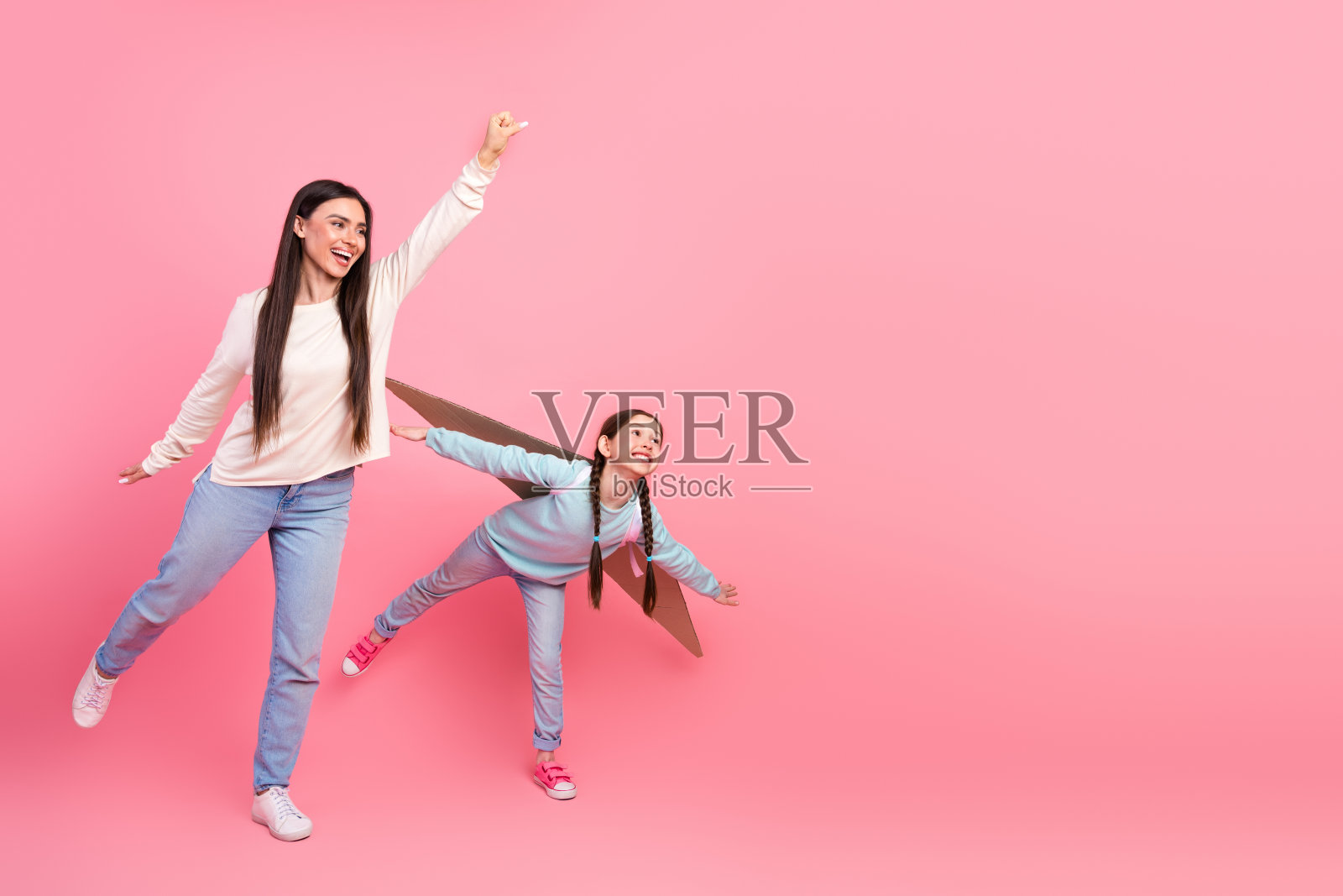 快乐的母亲和年轻女儿在粉色背景下玩纸板翅膀,展现家庭乐趣和想象力。照片摄影图片