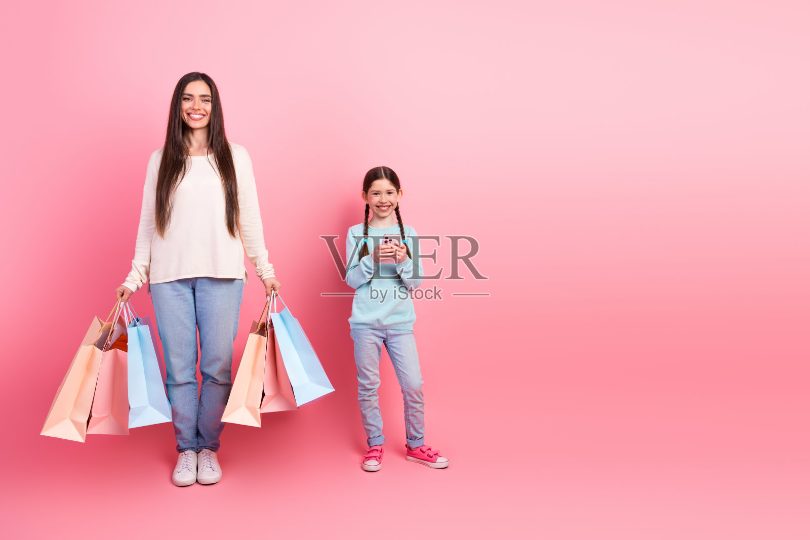 母女俩站在粉色背景前,手里拿着购物袋和智能手机。照片摄影图片
