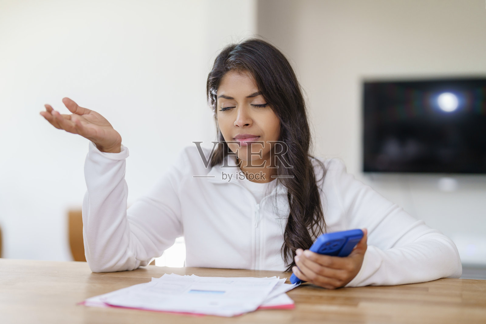 沮丧的女人坐在桌子旁,手里拿着手机,目光集中在文件上,周围是明亮的室内环境。照片摄影图片