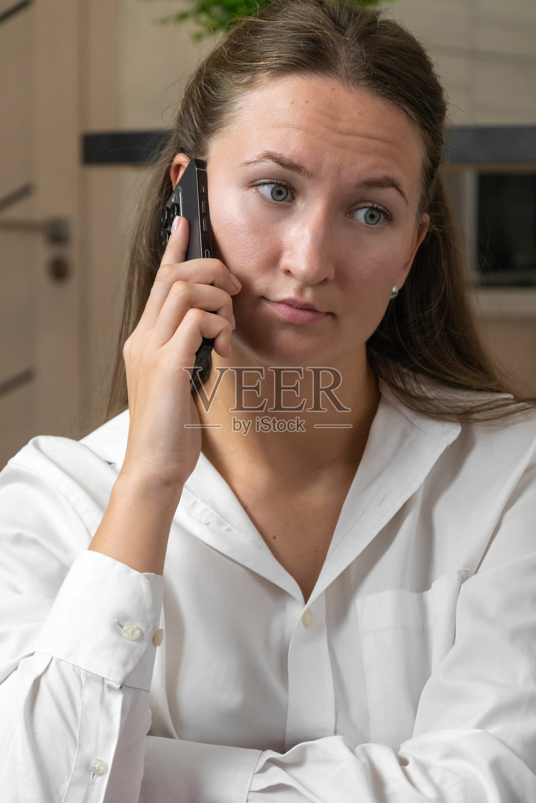 年轻女性穿着白衬衫,坐在沙发上用智能手机打电话,面露严肃神情,正在家中远程工作,听取重要消息。竖版照片照片摄影图片