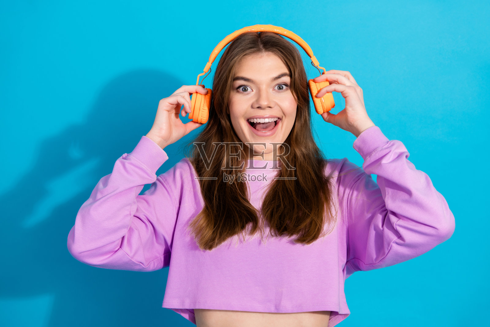 微笑的年轻女性穿着鲜艳的紫色卫衣,戴着橙色耳机,享受着音乐,背景是蓝色的。照片摄影图片