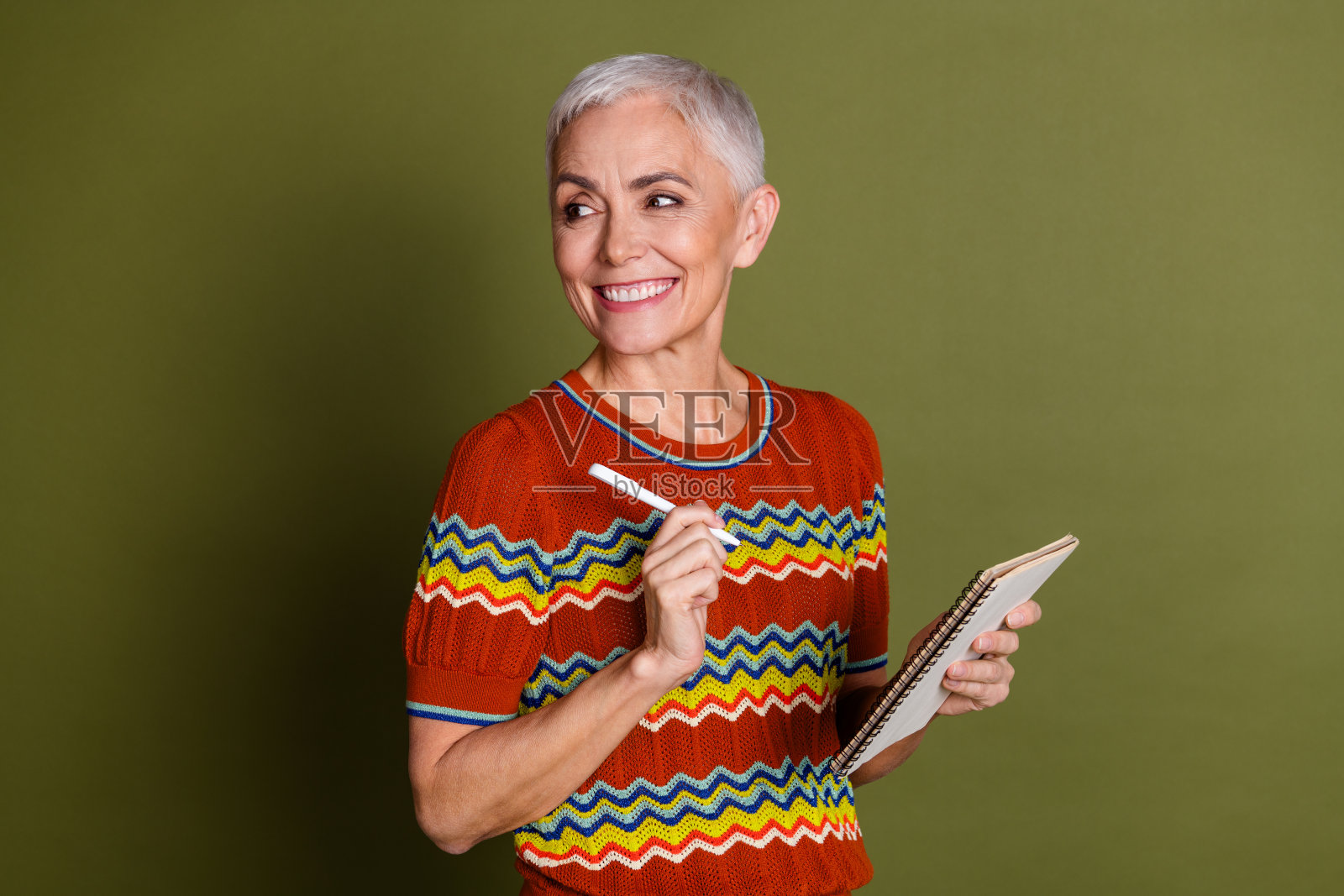 微笑的老年女性,穿着时尚的短发,身着多彩锯齿纹图案的休闲毛衣,背景为绿色,正在写笔记。照片摄影图片