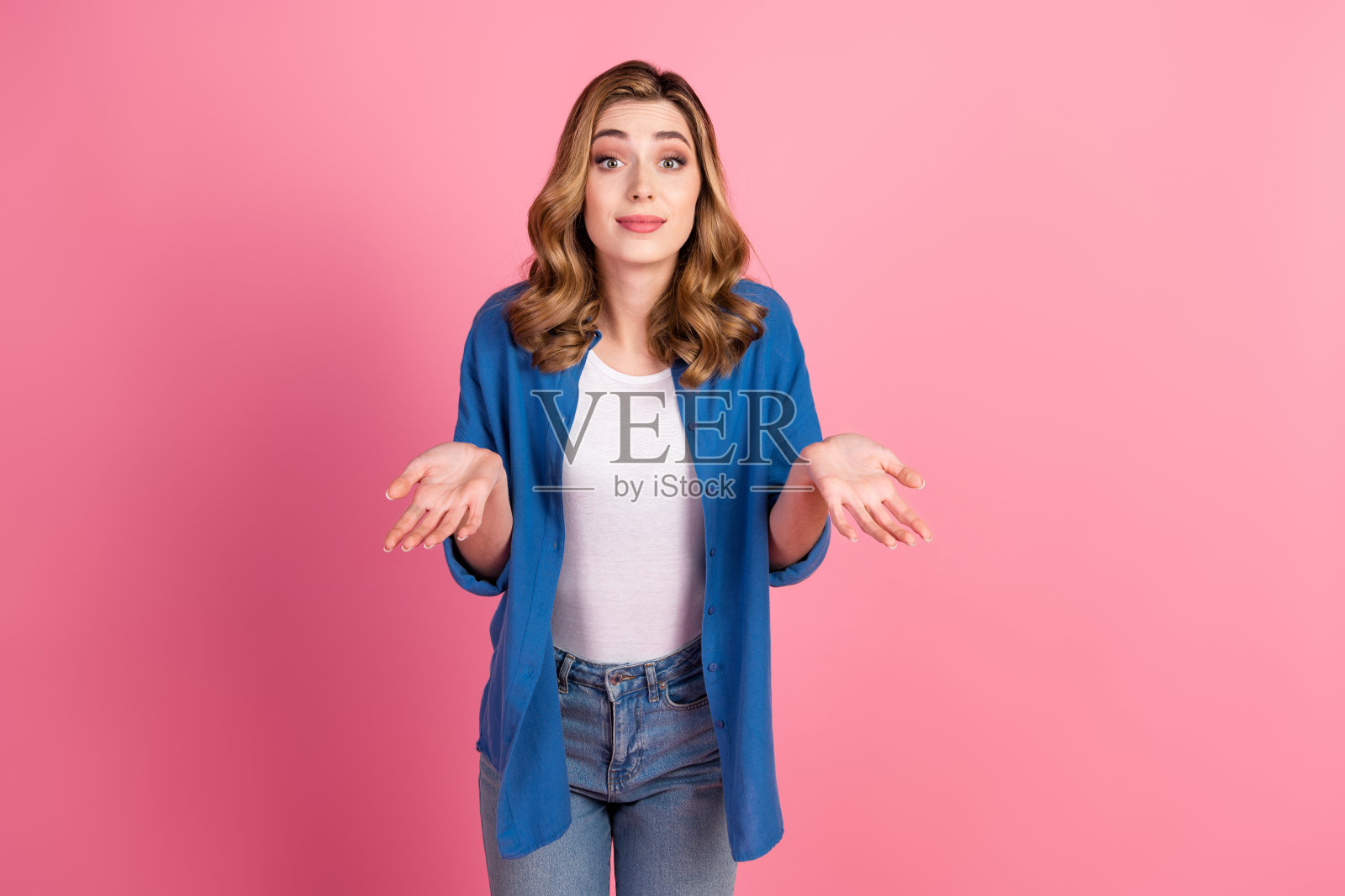 可爱的年轻女性,浅色头发,自信地在鲜艳的粉色背景前做手势,穿着休闲的蓝色衬衫和牛仔裤。照片摄影图片