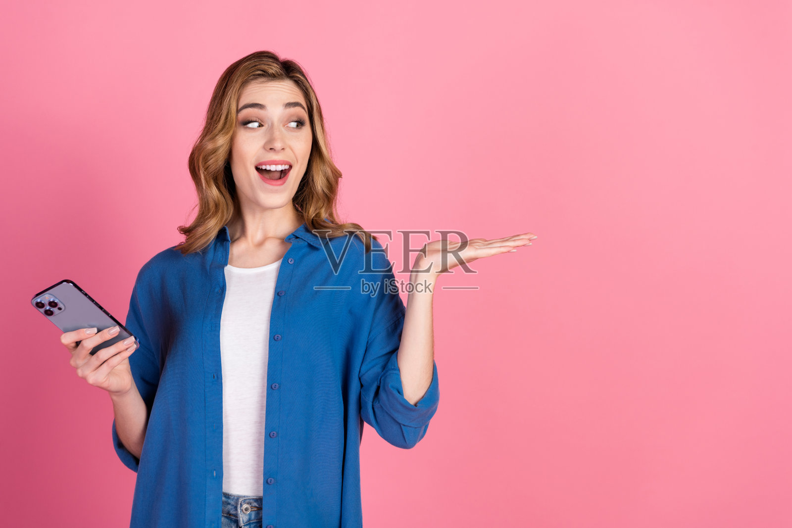 穿着蓝色衬衫的快乐女性手持智能手机,随意做手势,背景为鲜艳的粉色,适合生活方式推广。照片摄影图片