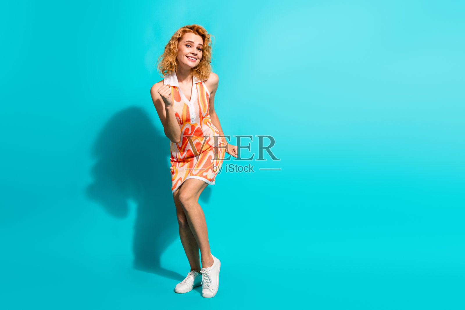 穿着色彩斑斓裙子的时尚年轻女性在明亮的青绿色背景前微笑并做手势,散发出快乐的气息照片摄影图片