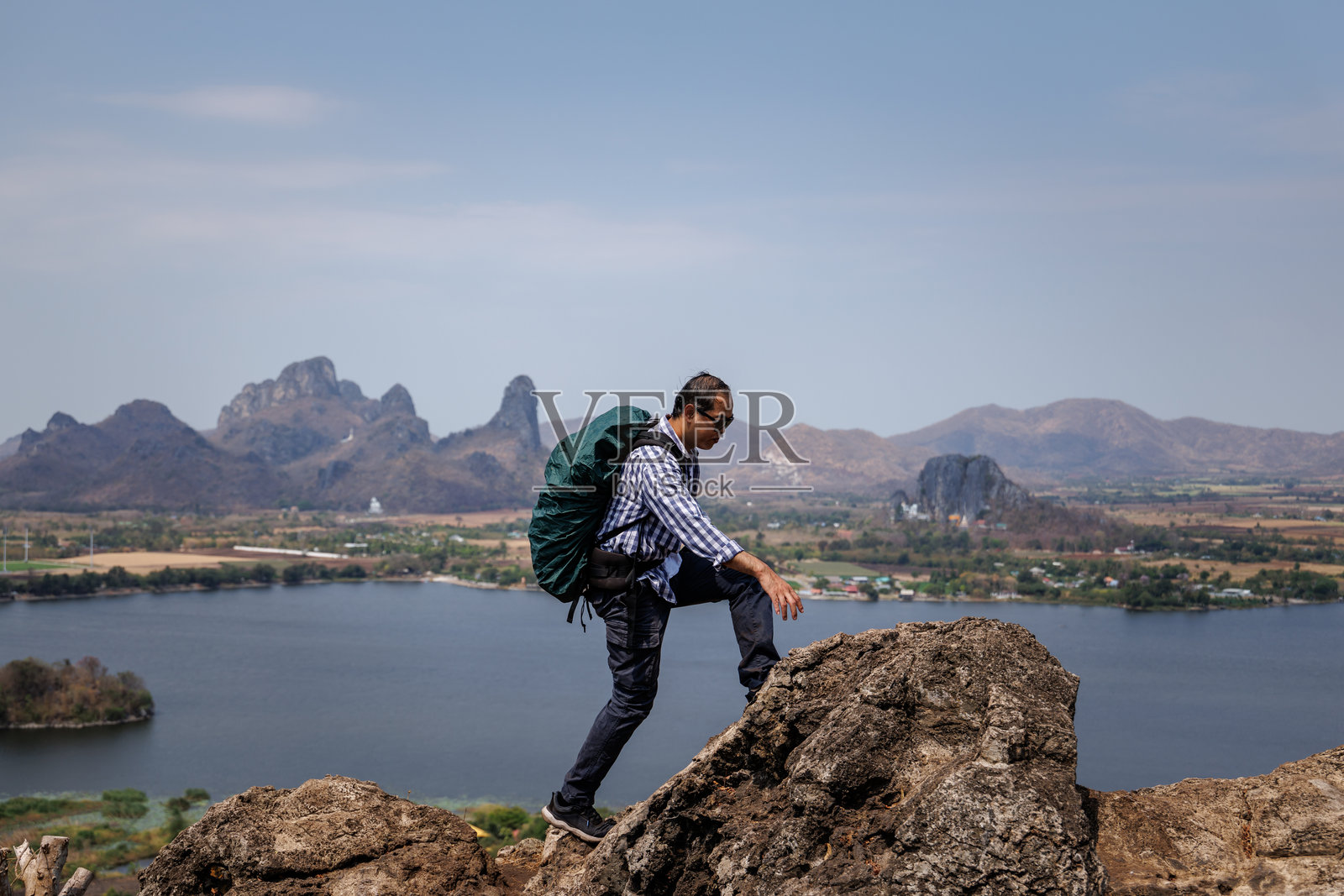 一名男性旅行者站在岩石山峰上,背着背包,凝视着下面的风景,风景中包括一个水库、一座小镇和远处的山脉。照片摄影图片