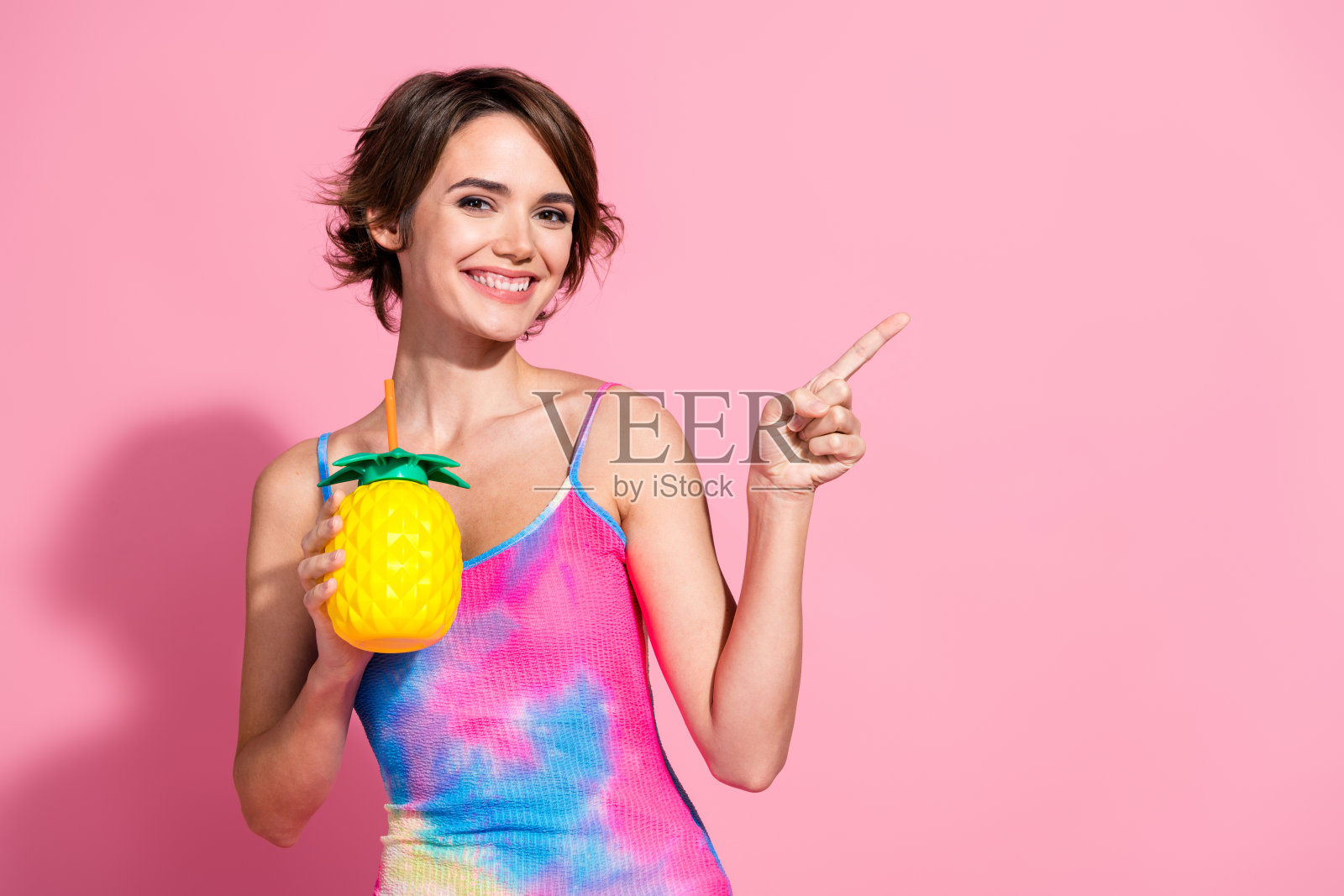 迷人的年轻女性穿着色彩缤纷的夏季连衣裙,手持菠萝杯,背景为粉色。照片摄影图片