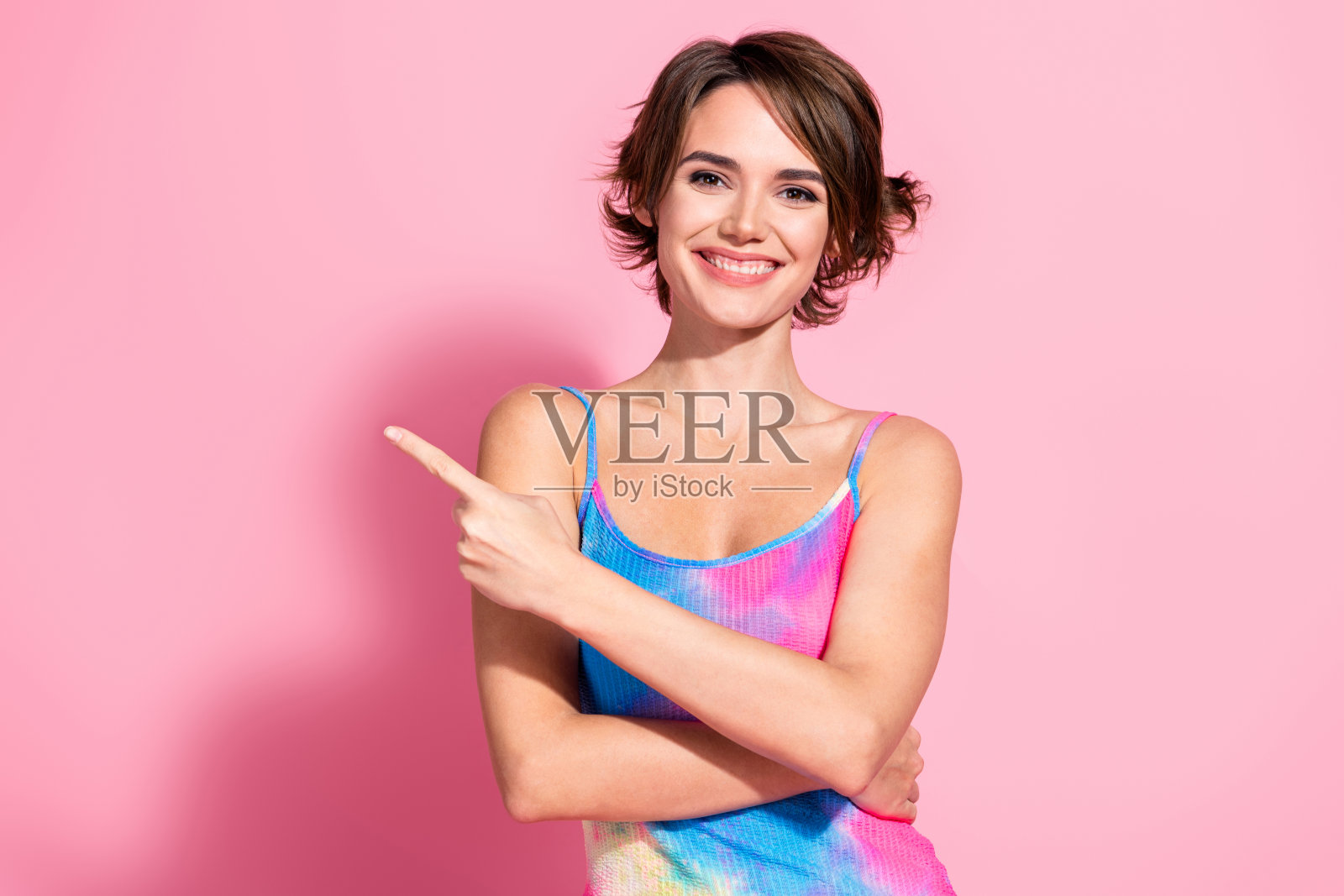 迷人的棕发女性穿着鲜艳的夏季连衣裙,在粉色背景前自信地做手势照片摄影图片