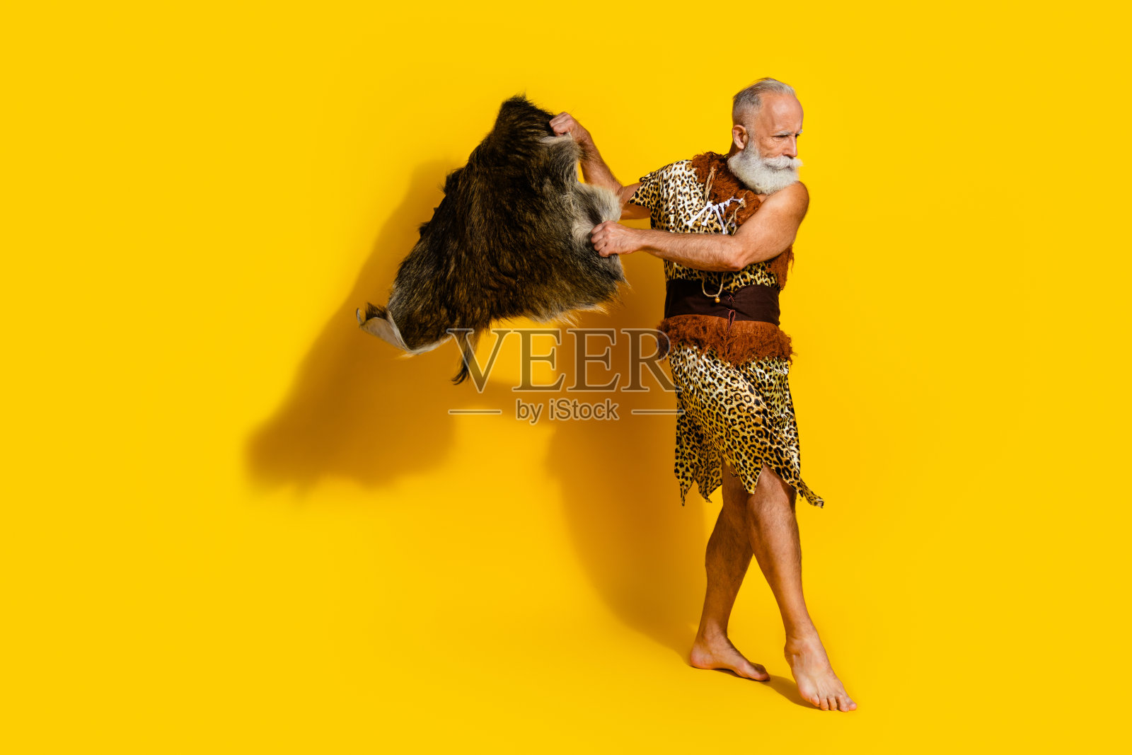 全身照,古老的穴居人穿着豹纹衣服,手持动物皮毛,背景为黄色。照片摄影图片