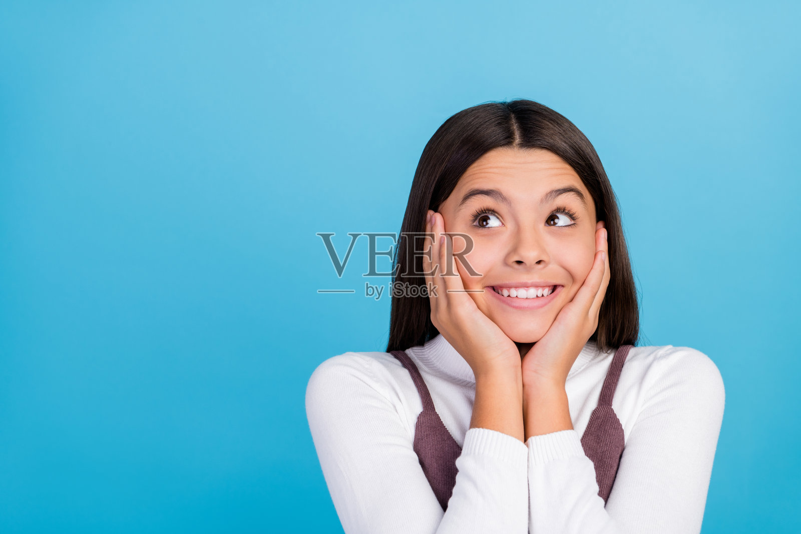 穿着棕色裙子的沉思梦幻学生女孩微笑着,双手托着脸颊,凝视着空旷的空间,背景为蓝色。照片摄影图片