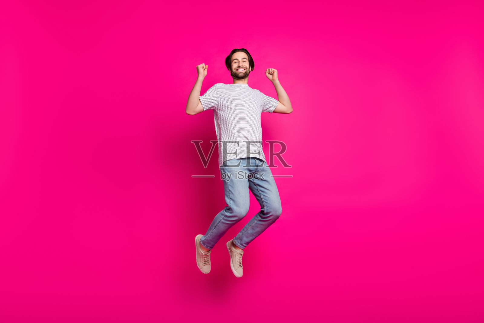 穿着条纹T恤、牛仔裤和运动鞋的惊讶胜利男孩在粉色背景下跳跃欢呼。照片摄影图片