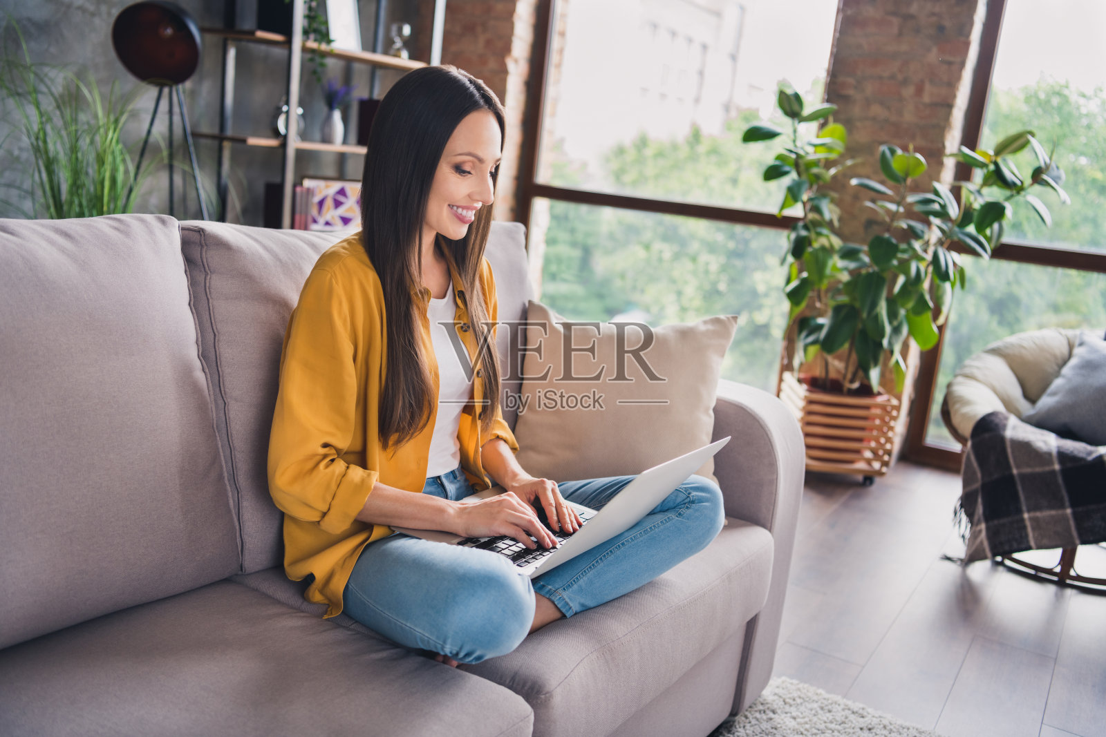 一位迷人的成熟的女性穿着黄色衬衫,坐在沙发上微笑着,正在室内的现代设备上打字。照片摄影图片