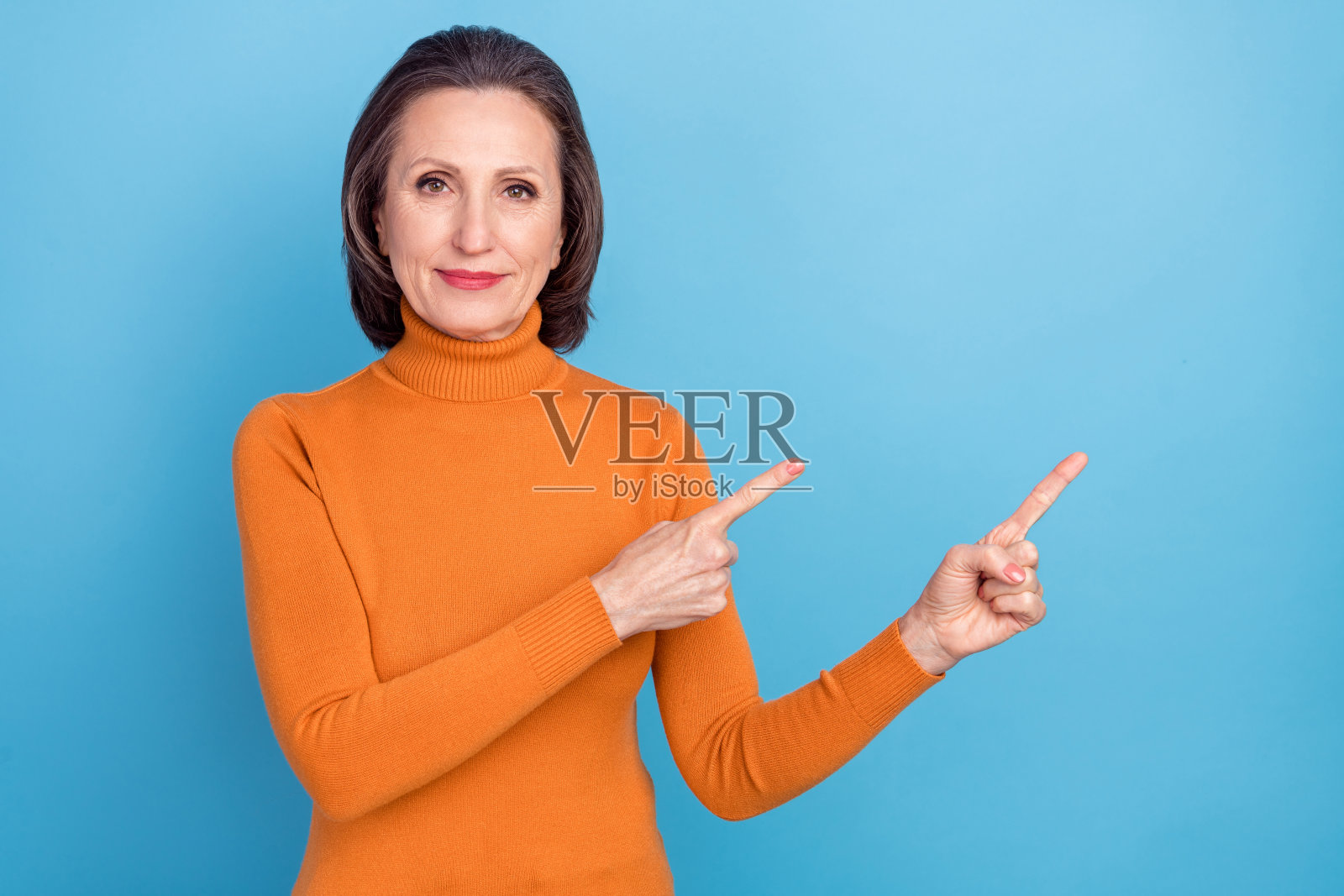 漂亮迷人的老年人用两根手指指向空白区域,提供新闻,背景为蓝色。照片摄影图片