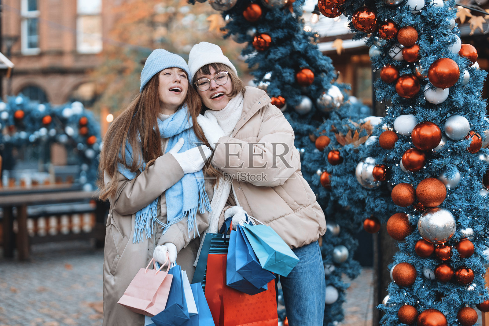 两个朋友穿着冬季服装,围绕着节日装饰享受购物的乐趣。照片摄影图片