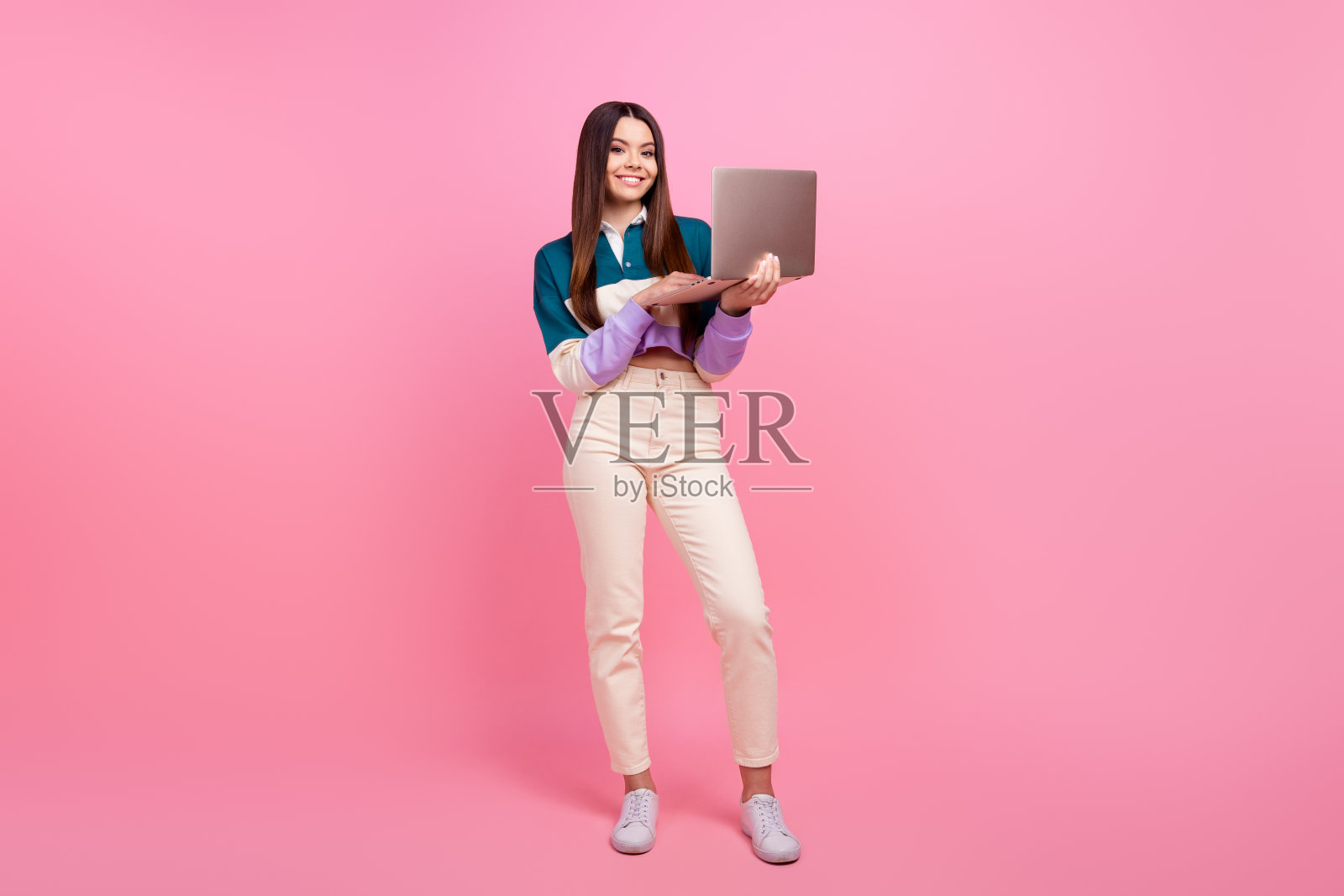 年轻女性手持笔记本电脑,微笑着站在粉色背景前,象征着现代生活方式照片摄影图片