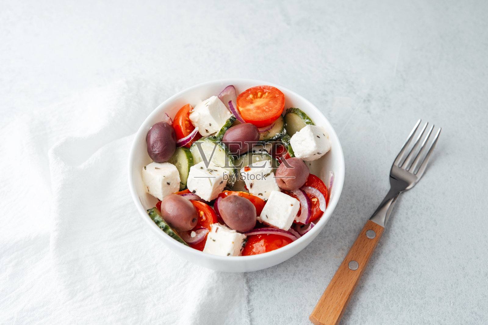 希腊沙拉,配有羊奶酪、橄榄、黄瓜和西红柿,放在白色碗中,旁边有叉子,背景为浅色。照片摄影图片