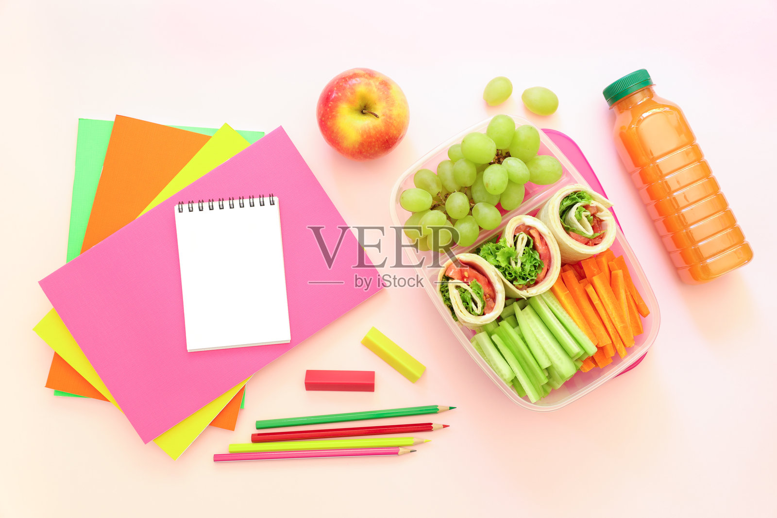 学校用品和午餐盒,里面有美味的卷饼、黄瓜、胡萝卜、葡萄、苹果和一瓶果汁,背景为浅粉色,回归校园的概念。照片摄影图片