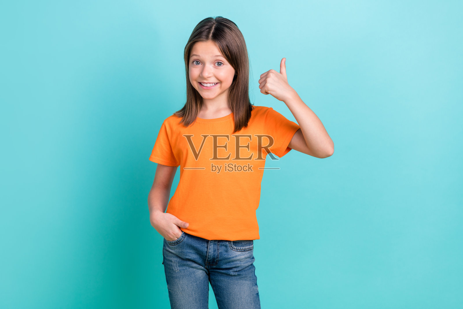 可爱的女孩竖起大拇指,穿着橙色T恤和牛仔裤,背景为青绿色。照片摄影图片