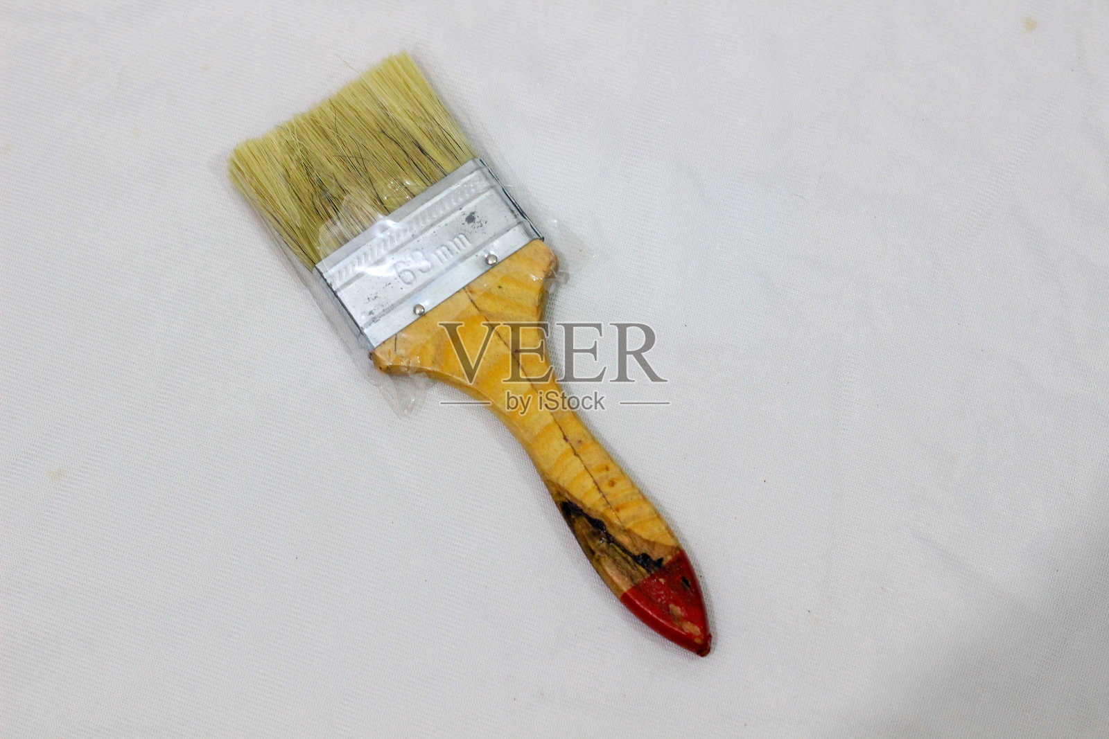 一把新的天然毛刷,刷毛覆盖着塑料,木柄带有红色涂漆的尖端,金属箍。刷子斜放在白色布料表面上。照片摄影图片