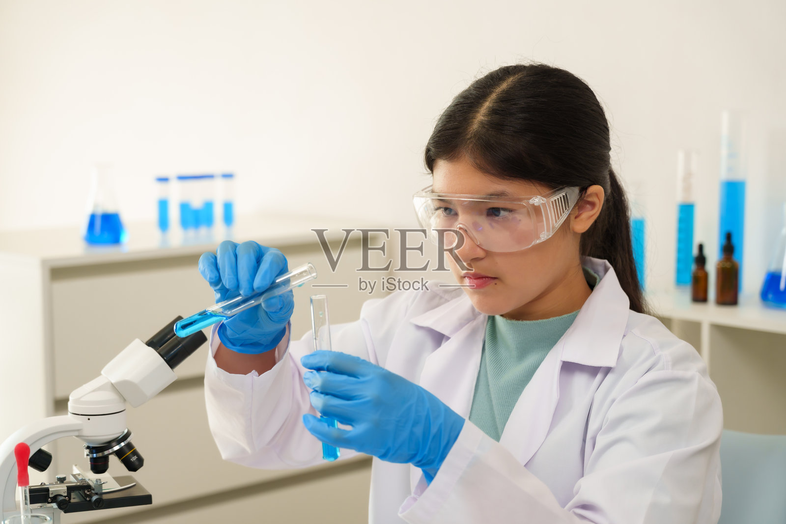 长发扎起的亚洲女孩穿着实验室外套和手套,专注地在学校科学课上进行化学实验,将液体倒入试管中。照片摄影图片