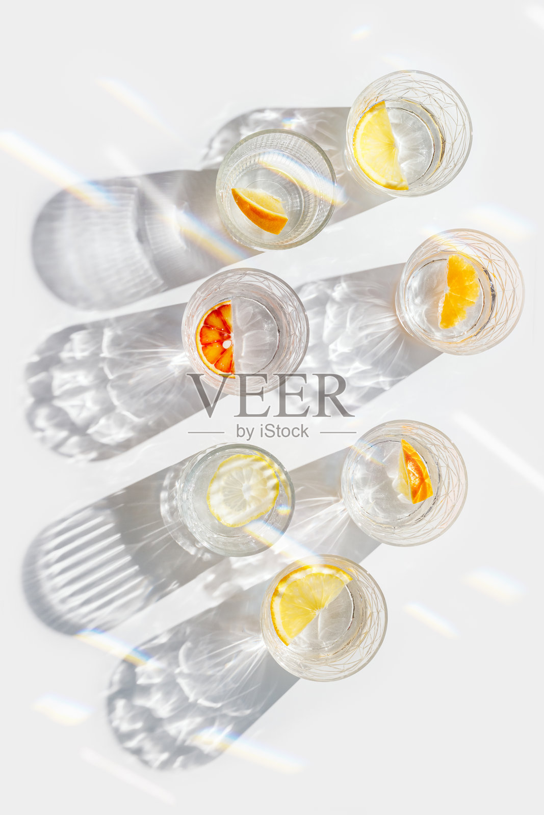 白色背景上,水中放有柑橘片的水晶杯,阳光下的彩虹反射,现代简约的构图,玻璃杯中装满水和柑橘水果,如柠檬和橙子。照片摄影图片