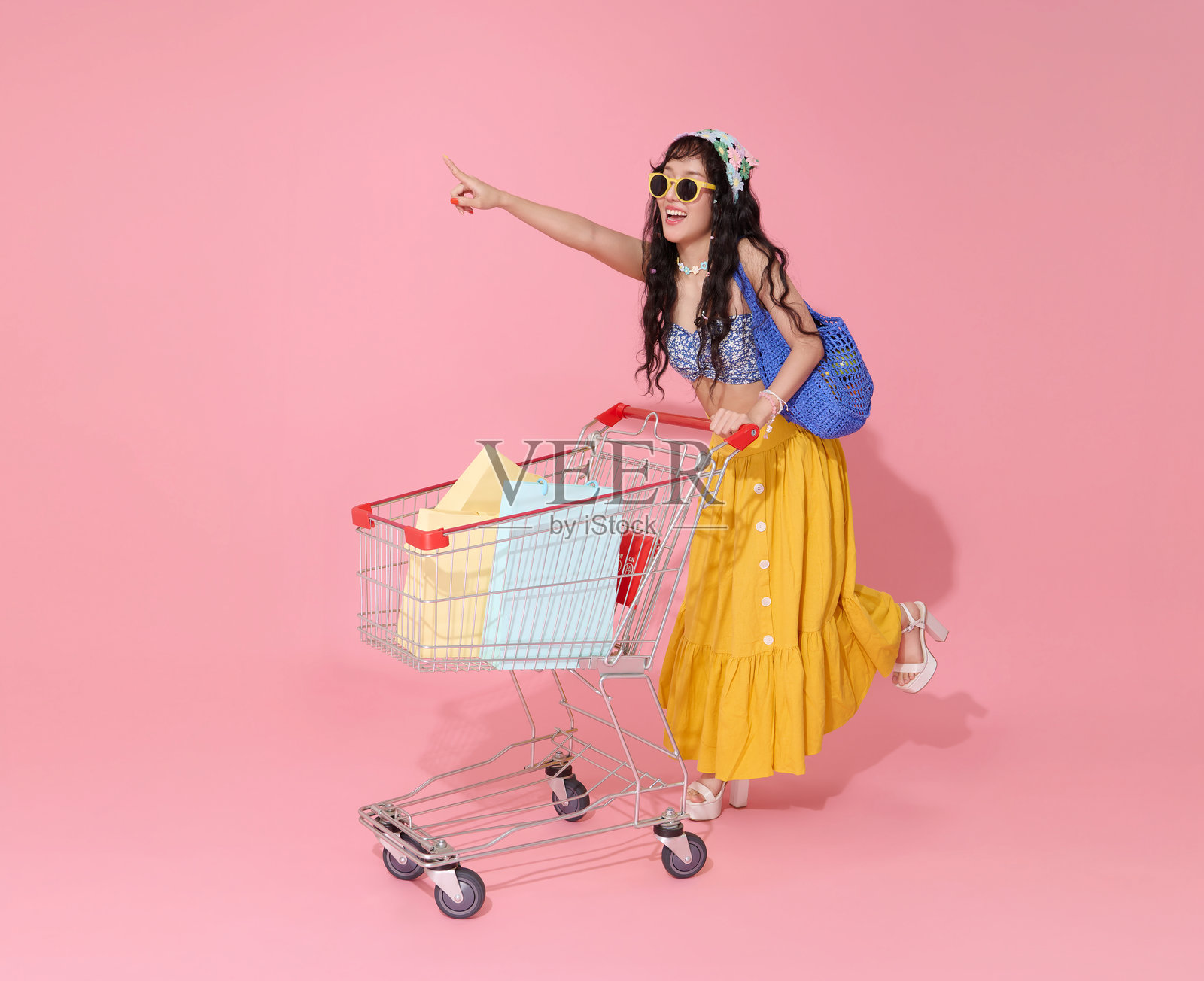 快乐微笑的年轻亚洲女性穿着夏季服装,推着购物车,手里提着夏季促销的购物袋,背景为粉色。照片摄影图片