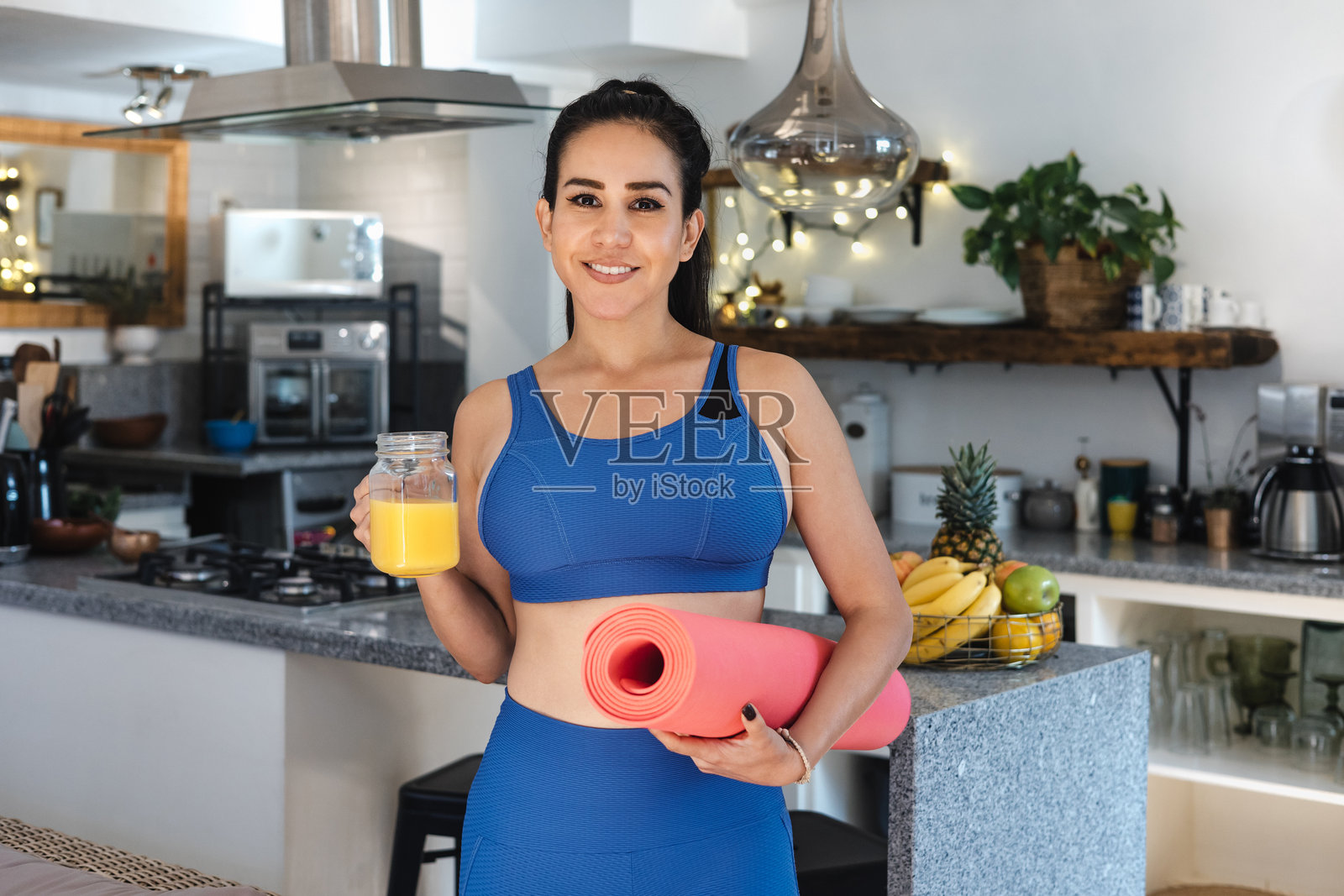 年轻拉丁女性在墨西哥的家中锻炼,喝着橙汁,手里拿着瑜伽垫,体现健康生活理念。照片摄影图片