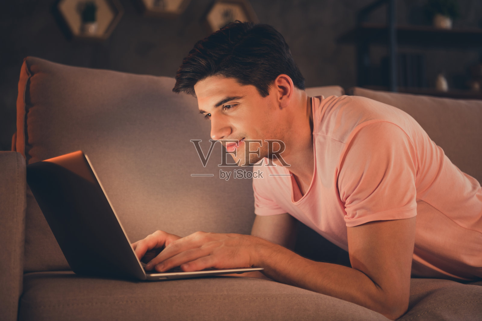 侧面视图的迷人、开朗、专注的男士躺在沙发上,使用笔记本电脑聊天和写邮件,室内环境。照片摄影图片